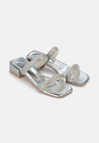 Sandali argentati con tacco quadrato, due cinturini luccicanti e un tacco a blocco metallico. Texture liscia e superficie riflettente.