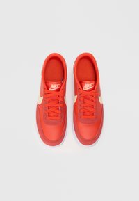 Červené tenisky Nike s hladkým koženým zvrškom, plochými šnúrkami a kontrastovým béžovým logom swoosh. Obsahujú polstrovaný interiér a bielu gumovú podrážku.