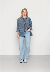 Casaco de camurça azul-claro com dois bolsos no peito, sobre uma blusa padrão azul-marinho, combinado com jeans de perna larga azul-claro e botas de tornozelo.