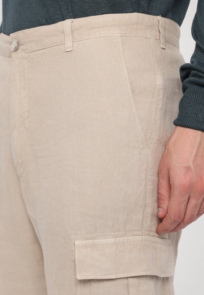 Lichtbeige katoenen cargo broek met zijzakken en een knoopsluiting. De stof lijkt textuur te hebben en is iets gekreukt.