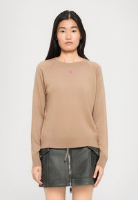 Kvinne iført en beige langermet genser med et lite rosa hjerte på brystet, og et svart skinnskjørt med frontglidelås og lommer.