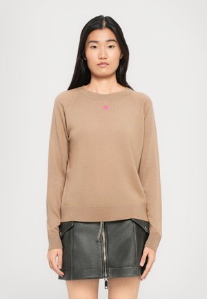 Femme portant un pull beige à manches longues avec un petit cœur rose sur la poitrine et une jupe en cuir noire avec fermeture éclair sur le devant et poches.