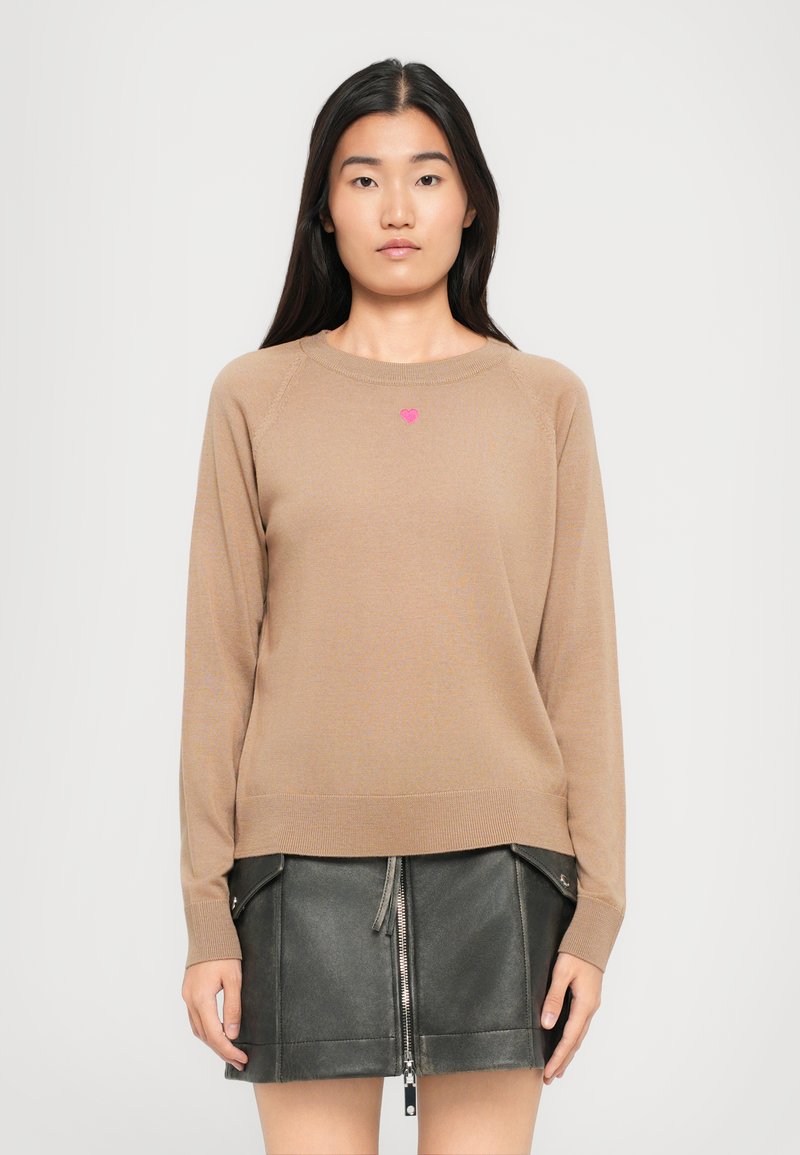 Vrouw in een beige langefotensweater met een klein roze hart op de borst en een zwarte leren rok met een rits aan de voorkant en zakken.