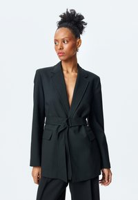 Blazer negro ajustado con escote en V profundo, cintura entallada gracias a un cinturón con una hebilla circular y dos bolsillos frontales. Textura de tejido suave.