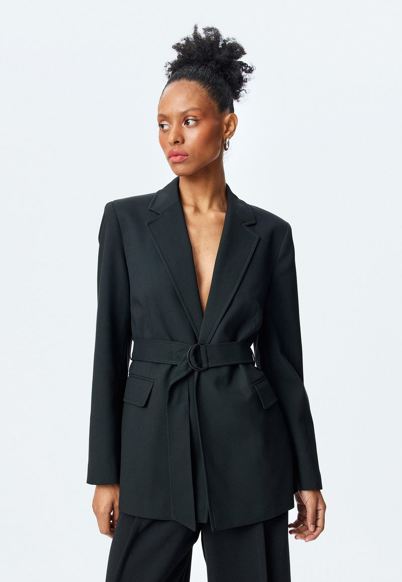 Blazer negro ajustado con escote en V profundo, cintura entallada gracias a un cinturón con una hebilla circular y dos bolsillos frontales. Textura de tejido suave.