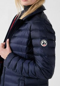 Vrouw in een marineblauwe gewatteerde jas met een ronde logo patch "Just Over The Top JOTT" op de bovenste mouw, terwijl ze het ritsgebied vasthoudt.