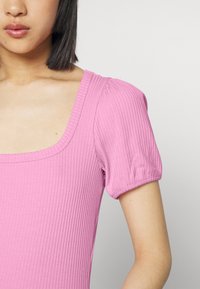Blusa ajustada em rosa pastel, com decote quadrado e mangas bufantes. O tecido tem uma textura suave com riscas horizontais.