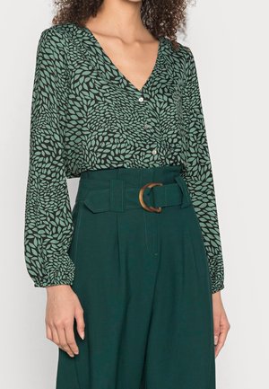 Blouse verte à motifs avec un col en V, des manches longues bouffantes et une fermeture à boutons, associée à un pantalon vert taille haute et une ceinture marron.