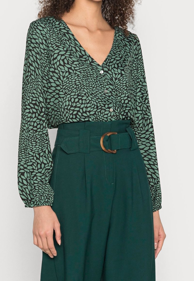 Blouse verte à motifs avec un col en V, des manches longues bouffantes et une fermeture à boutons, associée à un pantalon vert taille haute et une ceinture marron.