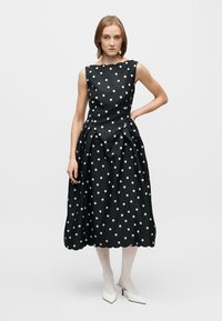 Svart ärmlös klänning med vita polka prickar, rynkad kjol och vågad kant. Modellen bär vita högklackade skor och strumpbyxor.