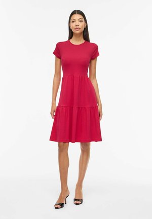 Ženska, oblečena v rdečo obleko s kratkimi rokavi do kolen, z oprijetim pasom in razširjenim krilom, obuta v črne sandale z odprtimi prsti, stoji na belem ozadju.