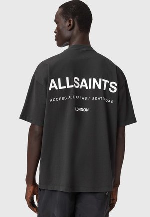 Homme portant un t-shirt noir oversize avec "ALLSAINTS Access All Areas / Backstage London" imprimé en blanc dans le dos.