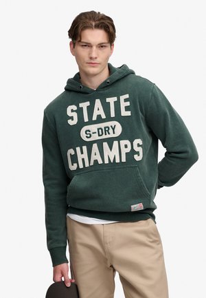 Ung mand iført en mørkegrøn hættetrøje med teksten "STATE S-DRY CHAMPS", beige bukser, holder en mørk kasket i den ene hånd.