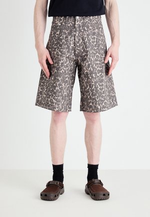 Homme portant un short marron et beige à motifs jusqu’aux genoux, des chaussettes noires et des chaussures marron sans lacets, debout devant un fond blanc.