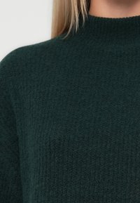 Tumši zaļš adīts džemperis ar ribotu tekstūru un augstu apkakli. Materiāls izskatās mīksts, norādot uz siltumu un komfortu.