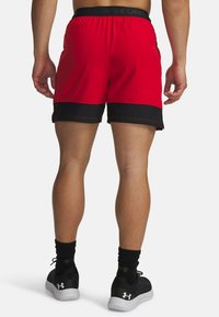 Pantalones cortos deportivos rojos con ribete negro, cinturilla elástica y detalles de malla. Combinados con zapatos y calcetines negros. Vista trasera.