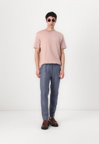 Hellpinkes Baumwoll-T-Shirt, lockerer Schnitt, kombiniert mit blauen plissierten Hosen. Das Modell trägt runde Sonnenbrillen und braune Slipper und steht vor einem weißen Hintergrund.