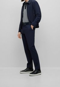 Veste bleu marine avec un polo gris à col, associée à un pantalon bleu marine ajusté et des baskets noires aux semelles blanches, avec une ceinture.