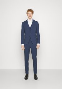 Marineblauer Anzug, bestehend aus einem taillierten Blazer und einer passenden Hose, mit einem weißen Hemd, schwarzen Schuhen und Knöpfen am Blazer.