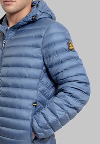 Giacca a piumino azzurro chiaro con cappuccio, caratterizzata da una texture trapuntata, tasche con cerniera e una toppa con logo sulla manica sinistra.