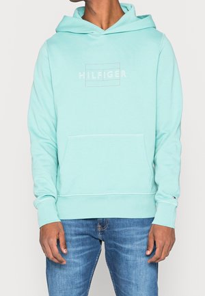 Mand iført lyseblå hættetrøje med teksten "HILFIGER" og frontlomme, kombineret med blå jeans, stående mod en ensfarvet baggrund.