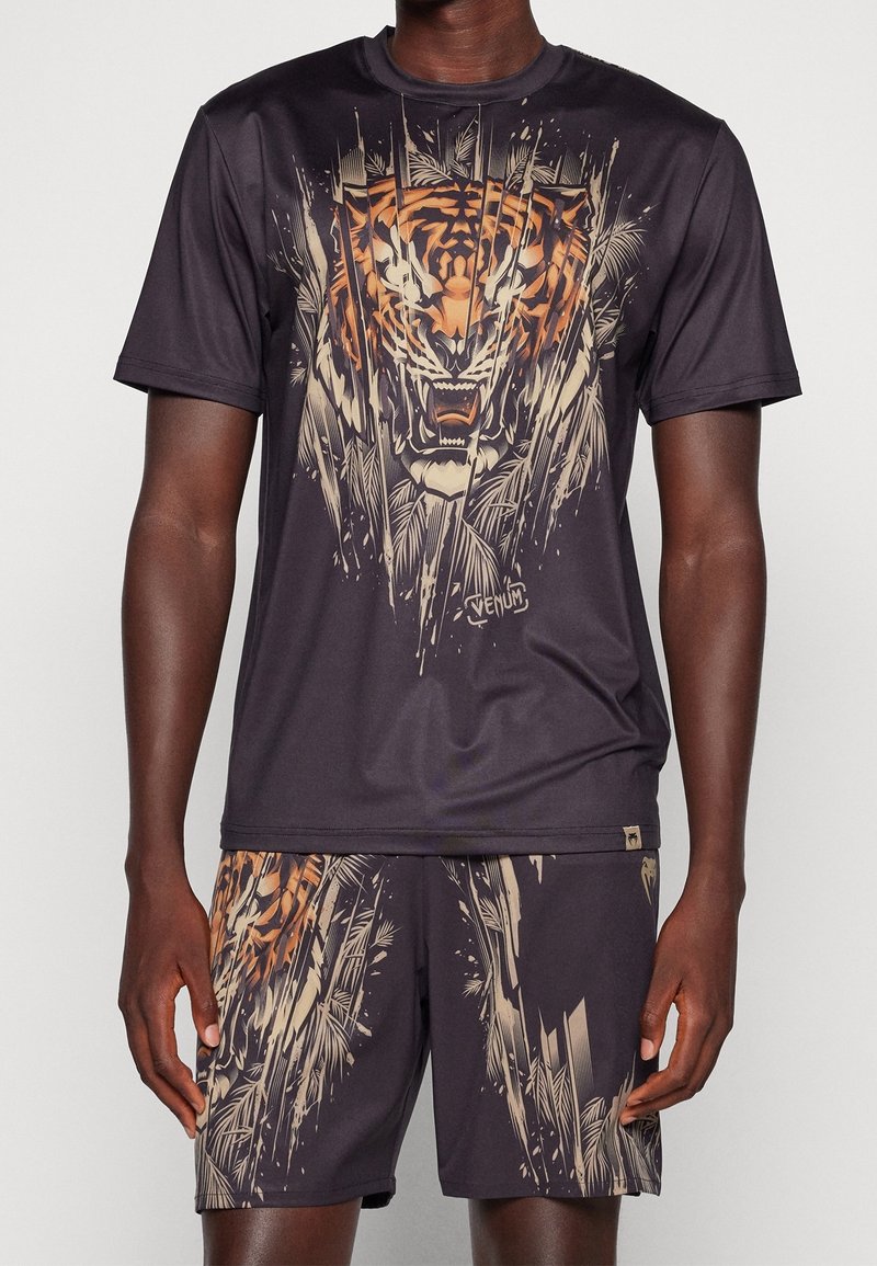 Man i svart sportset med stor rugande tigrer grafik i beige och orange på tröjan och shorts.