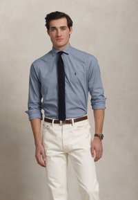 Lyseblå skjorte med oppbrettede ermer og en liten logo, kombinert med hvite jeans og et mørkt slips. Brun læderbelte og armbåndsur er synlige.