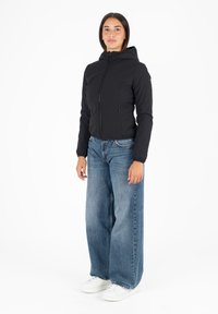 Giacca nera con cappuccio, dotata di zip frontale, due tasche laterali e polsini elasticizzati, abbinata a jeans in denim blu a gamba larga e sneakers bianche.