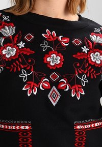 Czarny sweter z misternym haftem kwiatowym w kolorze czerwonym i białym na przodzie, z geometrycznymi akcentami i teksturowanym detalem ściegu u dołu.