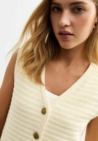 Cremfarbener gestrickter Pullover mit strukturiertem Muster, V-Ausschnitt, zwei goldfarbenen Knöpfen und taillierter Passform, übereinander getragen mit einem weißen Oberteil.