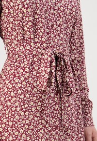 Rochie florală în burgundy, cu flori albe și galbene, având un cordon legat, închidere cu butoane în față și mâneci lungi cu manșete plisate.