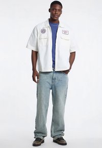 White short-sleeve shirt with two chest pockets and logos, worn over a blue t-shirt. Light blue jeans and black sneakers complete the outfit.