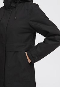 Veste noire imperméable avec capuche, cordons élastiques, poignets à bouton et poche avant, arborant un logo discret sur le côté inférieur.