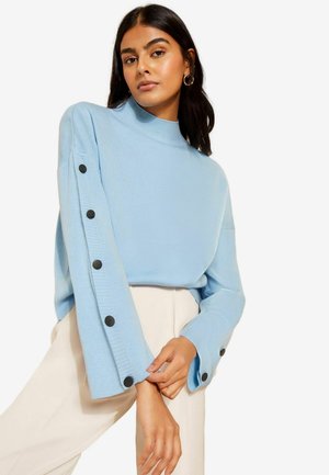Helles blaues, geripptes Sweatshirt mit hohem Kragen und langen Ärmeln, das schwarze Knopfakzente an den Bündchen aufweist. Dazu werden cremefarbene Hosen getragen.