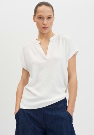 Frau mit zurückgebundenem Haar trägt ein strukturiertes weißes Kurzarmshirt mit Schlitz am Ausschnitt und dunkelblaue Hosen, Hände in den Taschen.