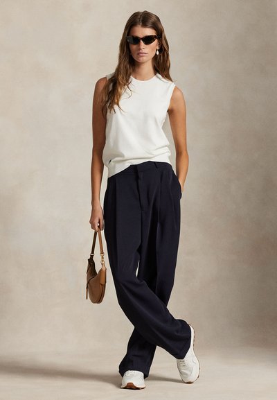 Top bianco senza maniche, pantaloni navy a gamba larga, sneakers bianche e borsa marrone. Modella con capelli lunghi e occhiali da sole scuri. Sfondo morbido.