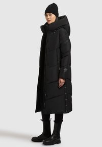 Abrigo tipo puffer negro con una silueta larga, capucha sobredimensionada y cierre con cremallera. Presenta secciones acolchadas y botones a presión en el costado.