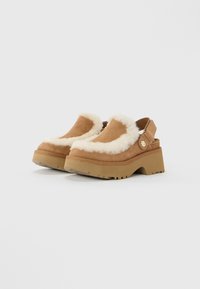 Brown suede platform klompen met dikke beige bontaccenten en verstelbare rugbandjes op een witte achtergrond.