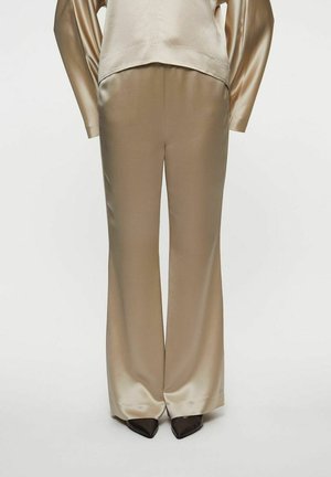 Beigen väriset satiinileveälahkeiset housut, yhdistetty samanlaiseen satiinitoppiin, mustien teräväkärkisten kenkien kanssa valkoisella taustalla.