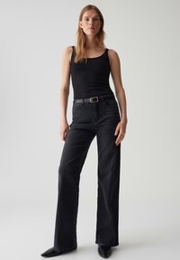 Femme debout devant un fond uni, portant un haut sans manches noir, un jean noir taille haute avec une ceinture et des chaussures noires à talons pointus.