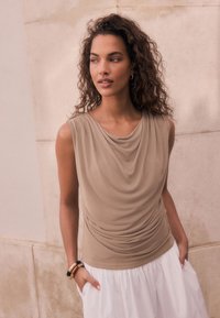 Haut sans manches beige avec encolure drapée et détails froncés à la taille, associé à une jupe blanche fluide. Texture de tissu lisse.