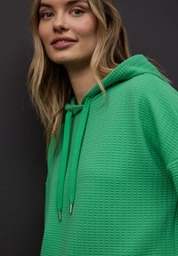 Femme aux cheveux châtain clair portant un sweat à capuche vert texturé avec des cordons, sur un fond gris foncé.