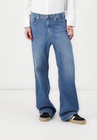 Vaaleansiniset, leveälahkeiset farkut, joissa on haalistunut pinta, viisi taskua ja nappisuljenta. Yhdistetty mustiin lenkkareihin, joissa on valkoisia yksityiskohtia.