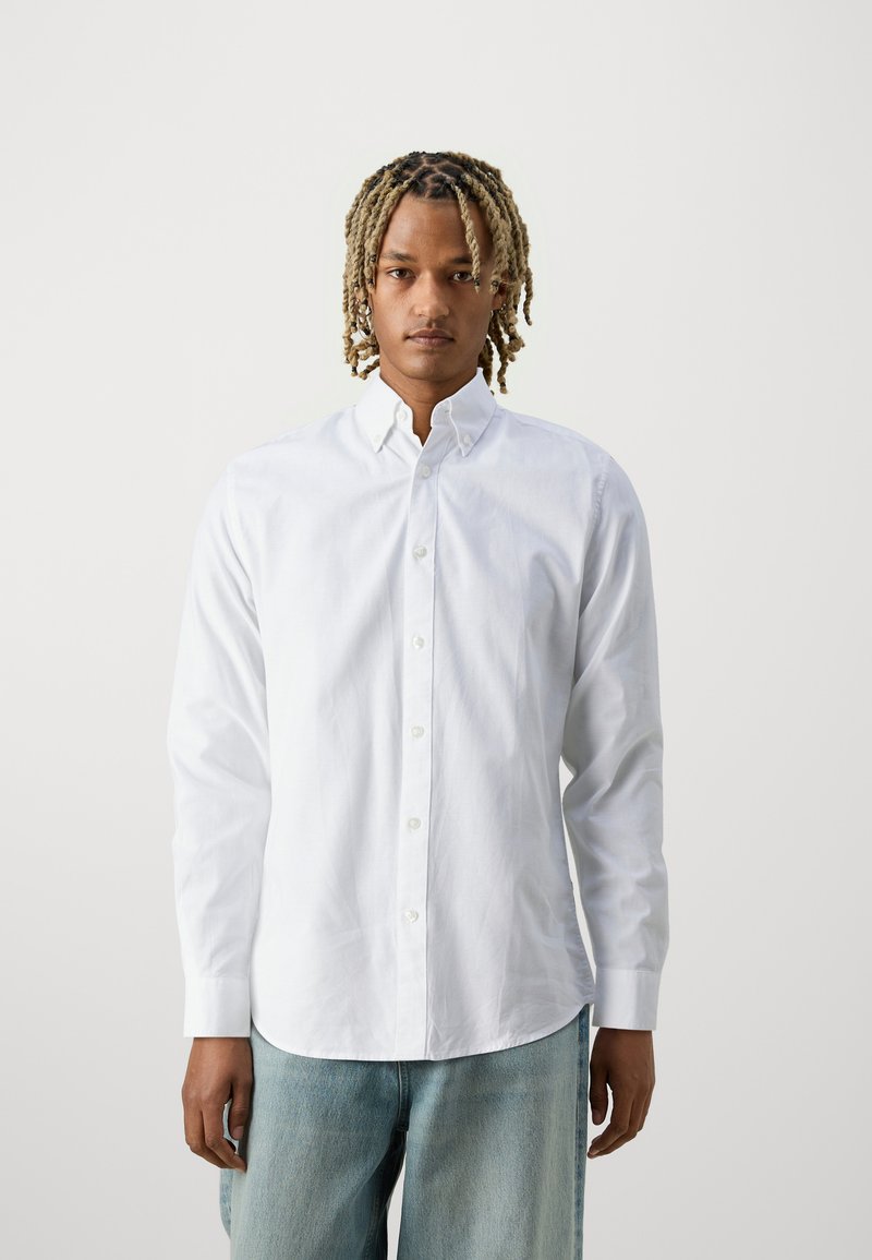 Camicia bianca con colletto standard e maniche lunghe, realizzata in tessuto liscio. Indossata con pantaloni in denim azzurro chiaro.