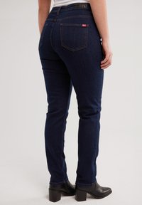 Isik, kes kannab tumedat sinist kitsaid teksapükse ja musta kontsaga pahkluusaapad, seisab lihtsa helerohelise tausta ees.