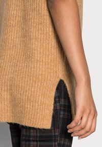 Hellbrauner, gerippter Pullover mit seitlichen Schlitzen. Weicher Stoff, lockere Passform. Kombiniert mit schwarzen karierten Hosen mit orangefarbenen Akzenten.
