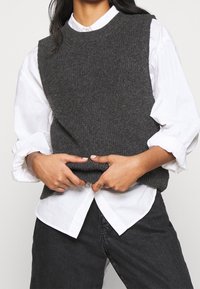 Chaleco de punto gris con escote redondo, tejido texturizado, llevado sobre una camisa blanca de botones, con mangas holgadas y jeans oscuros.