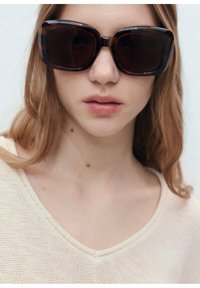 Tortoiseshell oversized sunglasses with dark lenses, featuring a thick frame and square shape, worn by a model with long, light brown hair.