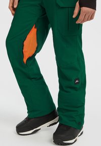 Groene skibroek met in oranje gemarkeerde mesh ventilatiepanelen, voorzien van meerdere zakken en een logo-opdruk bij de enkel. Zwarte schoenen.