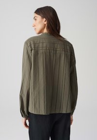 Camicia leggera verde oliva con righe verticali e schiena arricciata. Presenta maniche lunghe e una vestibilità rilassata. Dettaglio di tessuto testurizzato.